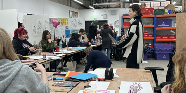 A group of happy students at L-shaped tables in an artistic studio surrounded by artwork and supplies listening to tutor