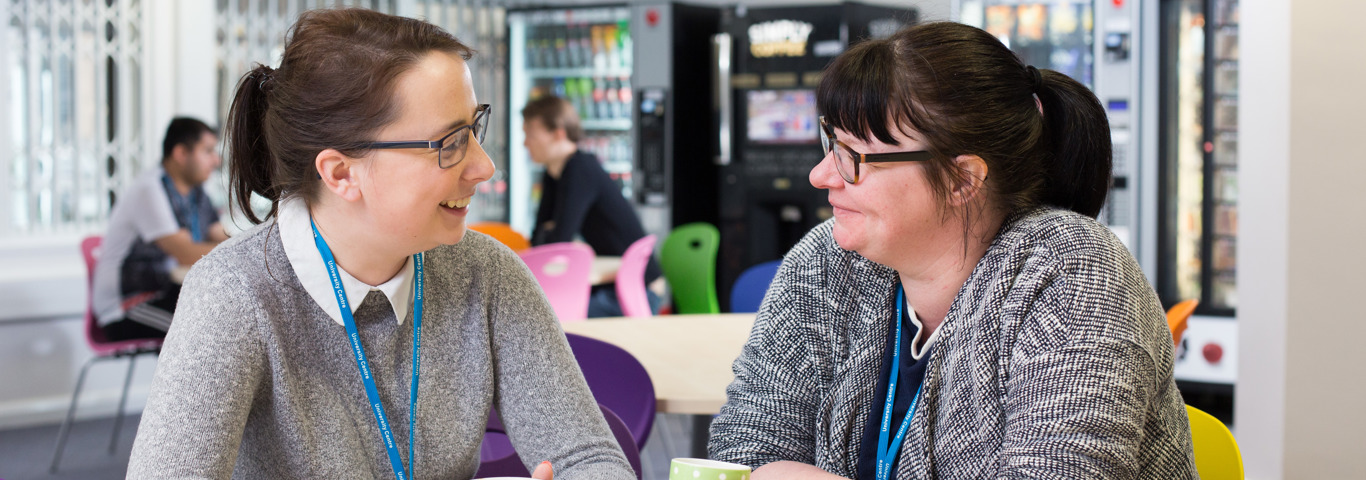 two staff members in a cafeteria holding cups of tea smiling