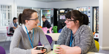 two staff members in a cafeteria holding cups of tea smiling